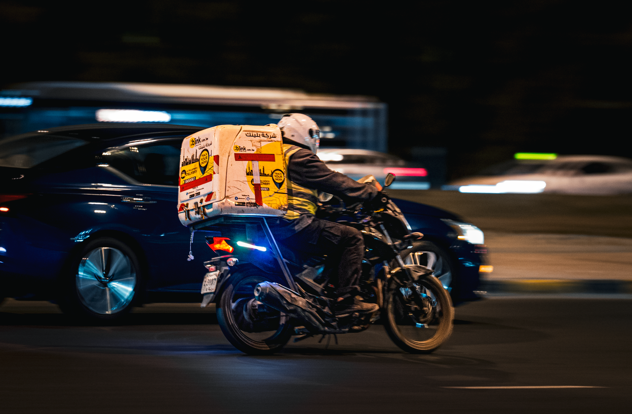 A delivery motorbike speeds through the streets of Kuwait City at night, showcasing fast-paced urban life.